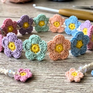 Op de afbeelding: Een handgemaakte gehaakte bloemenhoofdband met pastelkleurige bloemen en gele centra. De bloemen zijn gerangschikt rond een crèmekleurig koord, met extra bloemaccenten aan de uiteinden. Een haaknaald en schaar staan op de achtergrond.