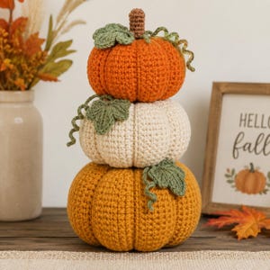 May include: A stack of three crocheted pumpkins in orange, cream, and mustard yellow. Each pumpkin features a brown stem, green crocheted leaves, and curly green vines. The pumpkins are stacked vertically, creating a decorative autumn-themed object.