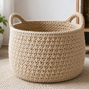 Puede incluir: Una cesta de almacenamiento redonda, de color beige, tejida a mano a ganchillo, con un patrón de puntos gruesos y texturizados y dos asas integradas en los lados opuestos. La cesta tiene una parte superior abierta y una base circular firme.
