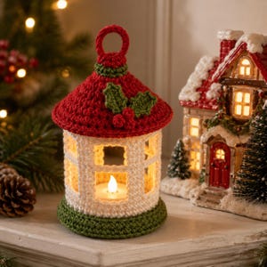 May include: A crocheted lantern featuring a white body with window-like cutouts, a red roof topped with a circular handle, and a green base. The roof is decorated with green holly leaves and red berries. A small LED tea light glows inside.