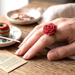 May include: A crochet ring featuring a vibrant red rose with green leaves on a thin green band. A brown paper tag nearby displays the text BLOOMING ROSE RING HANDMADE CROCHET with a rose illustration. A small ceramic dish holds yarn and a crochet hook.