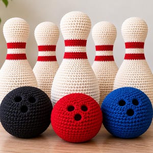 May include: A set of five cream-coloured crochet bowling pins with two horizontal red stripes around the neck, arranged behind three spherical crochet bowling balls in solid black, bright red, and deep blue, each featuring three finger holes.