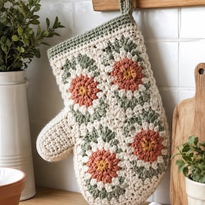 Könnte beinhalten: Ein gehäkelter Ofenhandschuh mit Granny-Square-Muster und Blumenmotiven in Creme, Salbeigrün und Terrakotta-Orange. Der Handschuh hat ein cremefarbenes Bündchen und eine passende Häkelschlaufe zum Aufhängen, gefertigt aus strukturiertem Garn.