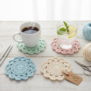May include: Four handmade crochet coasters in pastel colors: light green, pink, blue, and cream. A white mug with coffee and a glass of water with lemon and mint sit on two of the coasters. A tag reads "Handmade with Love."