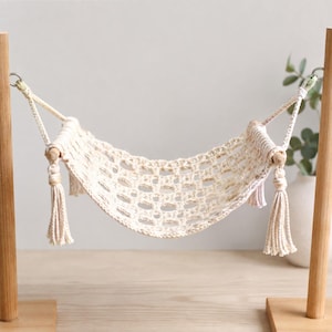 May include: A cream-colored macrame hammock suspended between two vertical wooden posts. The hammock features an open-weave knotted pattern with decorative tassels hanging from each side, attached to the wooden frame by metal rings.