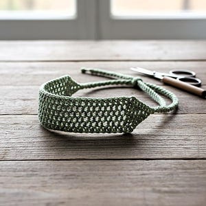 May include: A sage green crocheted headband with a textured, open weave design. The headband is on a wooden surface, with a pair of scissors in the background. The headband is adjustable and appears to be handmade.