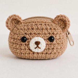Può includere: Un piccolo portamonete all'uncinetto color marrone chiaro a forma di testa d'orso. Presenta due orecchie arrotondate, due occhi a perla in plastica nera, un muso all'uncinetto bianco e un naso e una bocca ricamati in marrone scuro. Una cerniera beige è visibile sulla parte superiore.