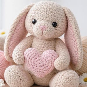 May include: A crocheted stuffed rabbit toy in a light beige color with long ears featuring soft pink inner linings. The rabbit has black plastic eyes, a stitched pink nose, and is holding a small, textured, heart-shaped pink crochet pillow.