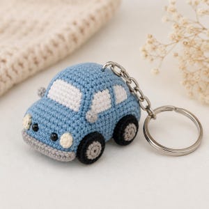 May include: A light blue crocheted car keychain featuring white windows, black and grey wheels, and small black bead headlights. The soft, stuffed vehicle is attached to a silver-toned metal split ring by a short chain, creating a compact accessory.
