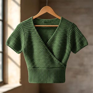 Puede incluir: Un top cruzado verde de manga corta hecho de tela de ganchillo. El top tiene cuello en V y dobladillo acanalado. La prenda se exhibe en una percha de madera sobre un fondo neutro.