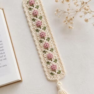 May include: A rectangular crochet bookmark in cream yarn featuring a repeating pattern of small pink rosebuds and green leaves. The edges are finished with a scalloped border, and a cream-colored yarn tassel is attached to the bottom end.