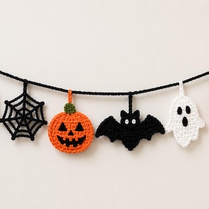 Op de afbeelding: Een Halloween-slinger met zes gehaakte ornamenten die aan een zwart garenkoord hangen. De ornamenten bestaan uit twee witte spoken, twee oranje pompoenen met groene steeltjes en zwarte gezichten, een zwart spinnenweb en een zwarte vleermuis met witte ogen.