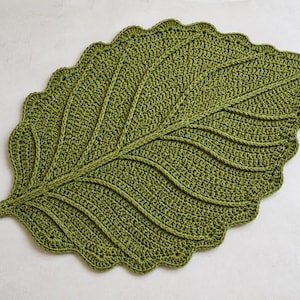 May include: A large, olive green crochet leaf mat featuring a detailed central vein and branching side veins. The edges of the leaf are scalloped, creating a decorative border. The entire piece is crafted with a consistent, tight stitch pattern.