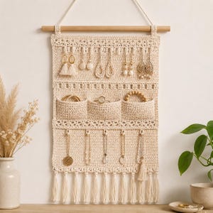 Puede incluir: Un organizador de joyas de pared tejido en crochet de color crema, suspendido de un listón de madera. Cuenta con múltiples bucles para colgar pendientes, tres bolsillos pequeños para anillos o pulseras, ganchos para collares y flecos inferiores.