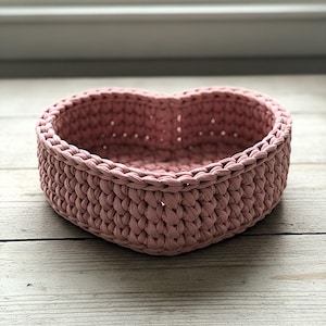 May include: A heart-shaped, dusty rose-colored crochet basket. The basket is made of thick, textured yarn and has a sturdy, woven appearance. The basket is sitting on a light-colored wooden surface.