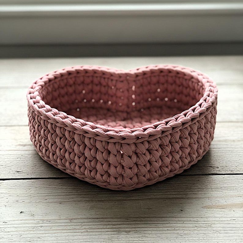Pode incluir: Uma cesta de croch&ecirc; em forma de cora&ccedil;&atilde;o, na cor rosa empoeirado. A cesta &eacute; feita de fio grosso e texturizado e tem uma apar&ecirc;ncia robusta e tecida. A cesta est&aacute; em uma superf&iacute;cie de madeira clara.