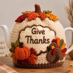 Può includere: Un copriteiera all'uncinetto color crema con la scritta Give Thanks ricamata in marrone. Il copriteiera è decorato con una zucca arancione, un tacchino marrone con piume colorate e foglie dai toni autunnali sulla parte superiore.