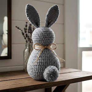Könnte beinhalten: Eine graue, gehäkelte Hasenfigur mit langen Ohren, rundem Körper und einem flauschigen Schwanz. Der Hase ist mit einer braunen Schnur gebunden. Er sitzt auf einer Holzoberfläche.