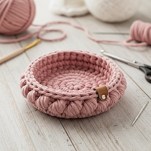 May include: A handmade, round, dusty rose crochet basket with a braided edge. A small brown leather tag is attached to the side. The basket is surrounded by yarn, a crochet hook, scissors, and a needle.