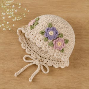 May include: A cream-colored crochet bonnet featuring a scalloped edge and two long ties. The bonnet is decorated with three small, hand-stitched flowers in shades of purple, pink, and light pink, each accented with green leaves and a yellow center.