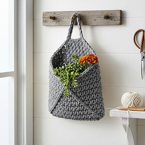 May include: A gray, crocheted wall pocket with a handle, hanging from a wooden plank. The pocket holds green foliage and an orange flower. A pair of scissors and a ball of yarn are visible in the background.