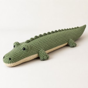 Pode incluir: Um brinquedo de crochê feito à mão em formato de crocodilo, com um dorso verde-sálvia texturizado e uma fileira de pequenas pontas triangulares ao longo da coluna. A parte inferior é de cor creme e o rosto possui dois olhos pretos.