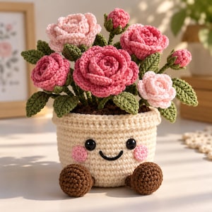 Könnte beinhalten: Eine gehäkelte Topfpflanze mit einem cremefarbenen Topf, der ein lächelndes Gesicht, rosa Wangen und zwei braune Füße hat. Der Topf enthält einen Strauß gehäkelter Rosen in hellen und dunklen Rosatönen, ergänzt durch grüne Blätter und Knospen.