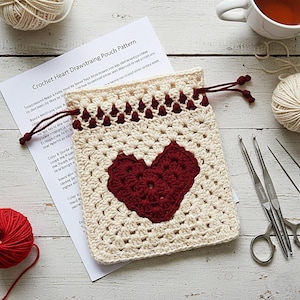 May include: A cream-colored crocheted pouch with a burgundy heart design. The pouch has a drawstring closure and is accompanied by crochet hooks, scissors, yarn, and a pattern titled "Crochet Heart Drawstring Pouch Pattern."