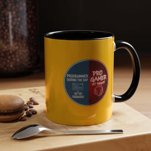 May include: A yellow ceramic mug with a black interior and handle. The mug has a design with the text "PROGRAMMER DURING THE DAY" and "PRO GAMER AT NIGHT" in a blue and red circle. A spoon and a pastry are on a wooden surface.