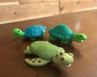 Peut inclure: Trois figurines de tortues en argile faites à la main, dans des tons de vert et de bleu. Les tortues ont des carapaces détaillées et des visages expressifs. Les figurines sont disposées sur une surface marron, mettant en valeur leurs motifs et couleurs uniques.