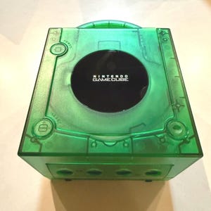 May include: A transparent green Nintendo GameCube console. The console has a black disc tray and the words "NINTENDO GAMECUBE" are printed on the top. The console has power and reset buttons, and several ports on the front.