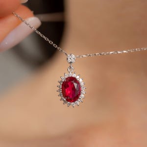 May include: A silver necklace featuring a large, oval-cut ruby pendant. The ruby is surrounded by a halo of small, sparkling diamonds. The necklace chain is delicate and the pendant is suspended from a small, diamond-studded bail.