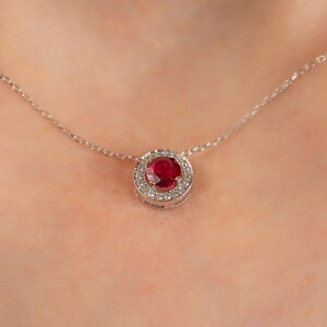 May include: A silver necklace featuring a round, deep red gemstone pendant encircled by a halo of smaller, clear gemstones. The pendant is suspended from a delicate silver chain, creating a classic and elegant look.
