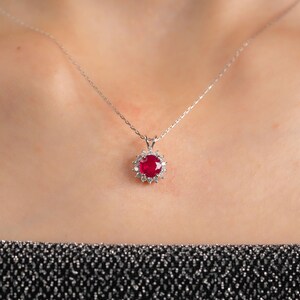 May include: A silver necklace with a pendant featuring a large, round, red gemstone surrounded by smaller, clear gemstones. The pendant hangs from a delicate silver chain. The necklace is displayed against a neutral background.