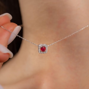 May include: A delicate silver necklace featuring a square pendant with a vibrant red gemstone at its center, surrounded by a halo of smaller, sparkling stones. The necklace is displayed against a skin tone background.