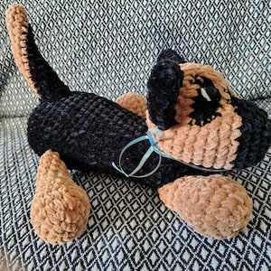 May include: A handmade crochet dog plushie in black and tan. The dog has floppy ears, a tail, and light blue ribbon around its neck. The plushie is lying on a patterned surface. The dog's paws are tan.