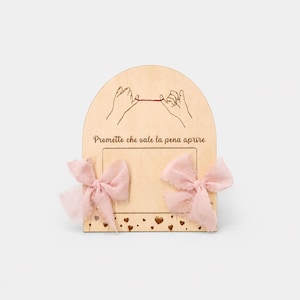 Può includere: Un ricordo in legno con un fiocco di nastro rosa su ogni lato. Il pezzo di legno ha una parte superiore ad arco con un disegno di due mani che tengono un filo rosso. Il testo "Prometto che vale la pena aprire" è sotto.