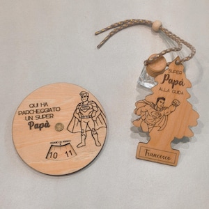Può includere: Due regali in legno a tema supereroi. Uno è un disco di parcheggio rotondo con testo e grafica di un supereroe. L'altro è un profumatore per auto a forma di albero con grafica di un supereroe e il nome "Francesco".