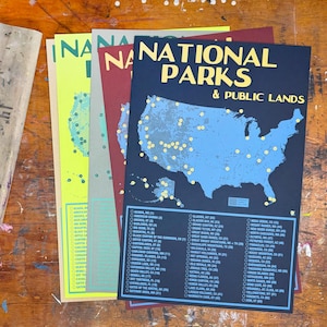May include: A stack of colorful posters with a map of the United States and the text "NATIONAL PARKS & PUBLIC LANDS." The posters are in shades of blue, yellow, and red. A wooden screen printing tool is on the left.