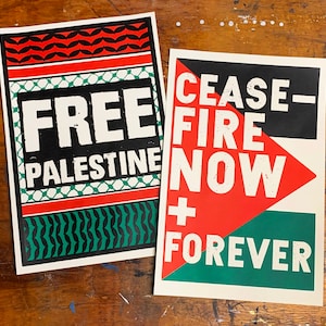 May include: Two protest signs with black, red, and green colors. The first sign says "FREE PALESTINE" with a geometric pattern. The second sign says "CEASE-FIRE NOW + FOREVER".