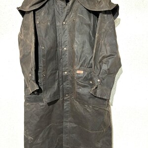 May include: A dark brown, full-length waxed cotton duster coat featuring a wide shoulder cape, a front button-down closure with metal snaps, long sleeves with adjustable cuffs, and two large front patch pockets with flaps.