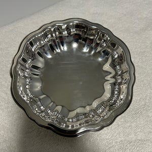 May include: A small, shiny silver-colored bowl with a scalloped edge. The bowl has a reflective surface, and the rim has a decorative, wavy design. The bowl is sitting on a white surface.