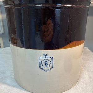 May include: A large, antique stoneware crock with a dark brown glazed top and a cream-colored base. The crock features a blue emblem with the number "6" and the letter "M". The crock is a vintage kitchenware item.