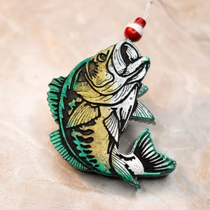May include: A decorative fish ornament with a green, yellow, and white color scheme. The fish has a detailed design and is attached to a red and white bobber with a string. The ornament is likely for fishing enthusiasts.