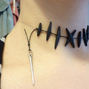 Puede incluir: Un collar gargantilla negro con un colgante de aguja. El collar est&aacute; hecho de cuentas negras y un cord&oacute;n negro. El colgante de aguja es de plata y tiene un peque&ntilde;o ojo en la parte superior.