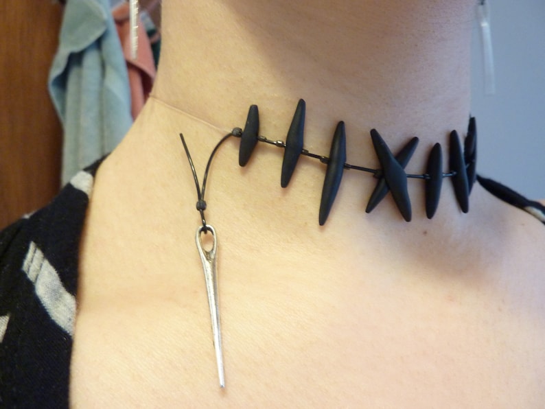 Puede incluir: Un collar gargantilla negro con un colgante de aguja. El collar est&aacute; hecho de cuentas negras y un cord&oacute;n negro. El colgante de aguja es de plata y tiene un peque&ntilde;o ojo en la parte superior.