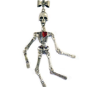 May include: A silver skeleton pendant necklace with a red heart and a bow at the top. The skeleton is posed with its arms outstretched and legs crossed.