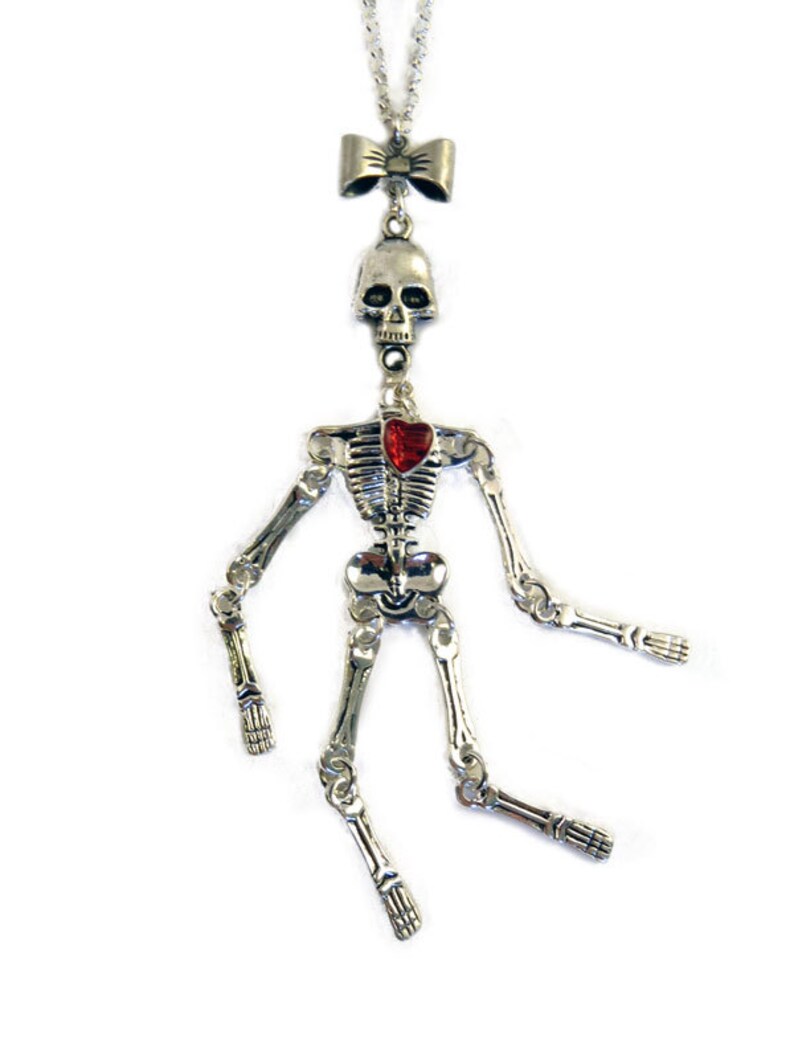 May include: A silver skeleton pendant necklace with a red heart and a bow at the top. The skeleton is posed with its arms outstretched and legs crossed.