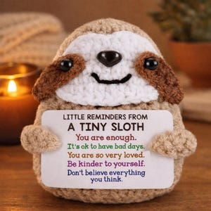 May include: A crocheted sloth figurine in shades of tan, brown, and white, holding a small card. The card reads "LITTLE REMINDERS FROM A TINY SLOTH" followed by inspirational phrases. The sloth has a smiling face and small black eyes.