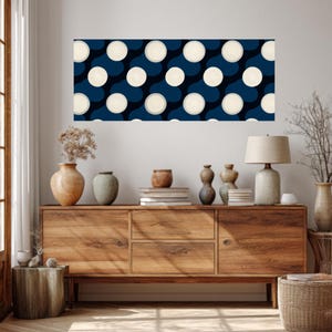 Op de afbeelding: Een rechthoekig kunstwerk met een herhalend patroon van grote, crèmekleurige cirkels op een donkerblauwe achtergrond. Het kunstwerk hangt boven een houten kast met diverse decoratieve vazen en een lamp.